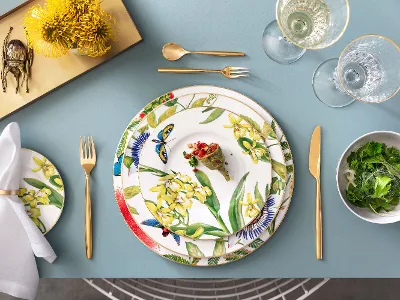 Amazonia - Florale Details und goldene Akzente Teller mit tropischen Blumenmotiven und goldenen Akzenten auf einem eleganten Tisch, umgeben von Besteck und Gläser.