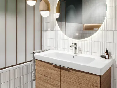 Architectura Waschbecken auf Holzwaschtisch mit rundem Spiegel Modernes Badezimmer mit großem runden Spiegel, einem Architectura Waschbecken auf einem Holzwaschtisch, hängenden Pendelleuchten und weiß gefliesten Wänden.