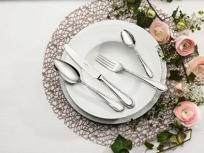 Posate Mademoiselle con rose colorate in tonalità pastello Un set di piatti bianchi di Villeroy & Boch Mademoiselle con coltello, forchetta e due cucchiai è elegantemente disposto su un centrino di pizzo, mentre fiori bianchi e rosa abbelliscono la tavola.