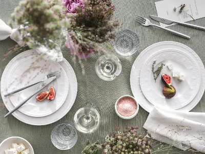 Cellini Geschirr für 2 Personen auf gedecktem Tisch Auf einem wunderschön gedeckten Tisch stehen Cellini-Teller von Villeroy & Boch mit Feigen, Käse und Kräutern. Gläser, Besteck, zarte Blumenarrangements und eine Schale mit Salz auf einer grünen Tischdecke runden das Ambiente ab.