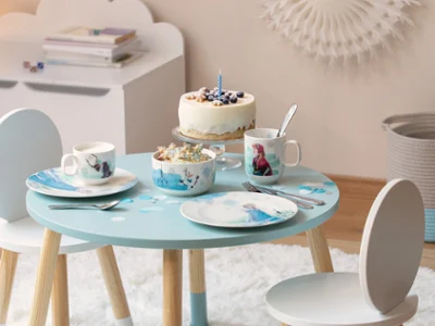 Tovagliato Disney Frozen Un piccolo tavolo blu, apparecchiato con stoviglie Frozen, tazze e una torta con mirtilli e una singola candela blu.