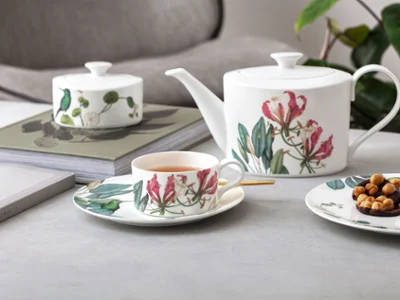 Avarua tea set on a table A teapot, a cup and a plate with snacks in a floral pattern are arranged on a table next to books and a plant in the background.