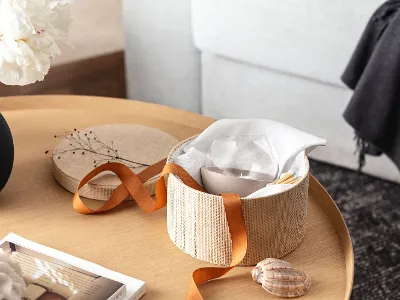 Une corbeille ronde tressée avec un nœud orange, dans lequel se trouve un photophore NewMoon Home, est posé sur une table en bois à côté d’un vase Collier noir.