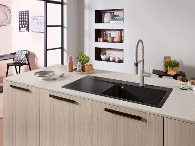 Subway Style Küchenspüle in heller Kücheninsel mit Armatur Eine moderne Küche mit einer schwarzen Spüle auf einer Kücheninsel aus hellem Holz, einem Wasserhahn, einer Pflanze und verschiedenen Gegenständen.