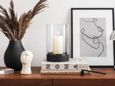 Manufacture Windlicht auf modernem Highboard Ein modernes Highboard mit Dekorationsgegenständen, darunter eine Vase mit getrockneten Pflanzen, ein schwarzes Manufacture Windlicht, eine Skulptur, gestapelte Bücher und ein gerahmtes abstraktes Kunstwerk.