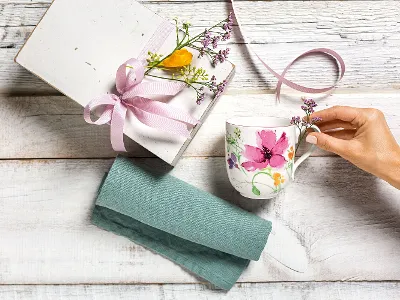 Eine Hand greift nach einem Villeroy & Boch Mariefleur-Becher neben einer Geschenkbox mit einer rosa Schleife und Blumen, die auf einer Holzoberfläche mit einem blaugrünen Tuch steht.
