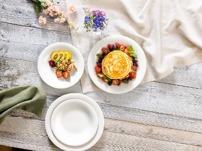 New Cottage Teller mit Pancakes Ein Stapel Pfannkuchen mit Beeren wird auf einem Teller von Villeroy & Boch New Cottage serviert, auf einem anderen Teller gibt es geschnittenes Obst. Der Holztisch ist mit Blumen und einer drapierten Tischdecke geschmückt.