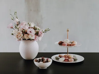 Eine weiße Vase mit rosa Blumen, ein mehrstöckiges Tablett mit Desserts und eine Schale mit Pralinen auf einem dunklen Tisch.