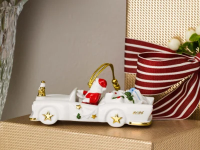 Cadillac 2026 gift A ceramic Christmas car with presents is sitting on a gift wrapped in gold, next to a vase of red and white flowers.