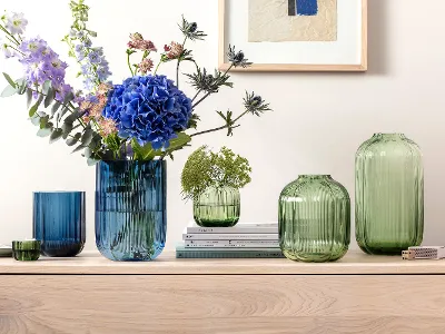 Ulike grønne og blå glassvaser på en treoverflate Ulike grønne og blå glassvaser på en treoverflate, noen med blomster og planter. På veggen bak henger et innrammet kunstverk.
