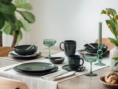 Une table à manger dressée avec Lave vert Une table à manger avec de la vaisselle verte, des verres, des couverts, une bougie et un vase avec des fleurs jaunes.