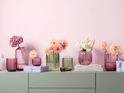 Green and pink Fleur glass vases on a green cabinet Various pastel-colored glass vases from Fleur stand on a green cabinet with stacked books and small gift boxes in front of a pink wall.
