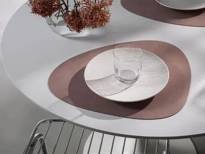Ein Glas und ein Teller stehen auf einem abstrakten braunen Tischset von Villeroy & Boch Lifestyle Accessoires auf einem weißen Tisch, im Hintergrund stehen ein Metallstuhl und eine Vase mit Trockenblumen.
