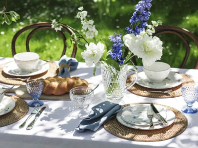 Vieux Luxembourg dinnerware on an outdoor table A garden table is set with “Alt Luxemburg” floral porcelain from Villeroy & Boch, wicker placemats, blue-tinted glass dinnerware, a crystal jug with blue and white flowers, and a loaf of bread.