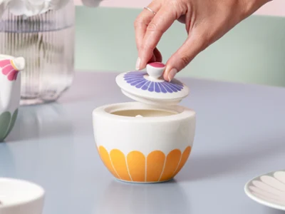 Fleur sugar bowl with yellow and purple patterns on a light blue table A hand lifts the lid of the Fleur ceramic sugar bowl with yellow and purple patterns on a light blue table.