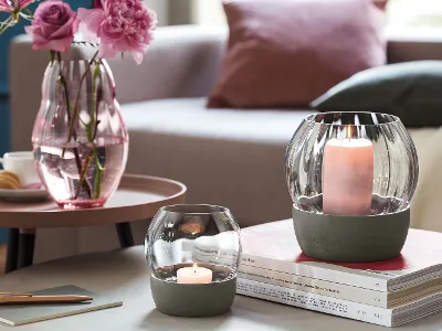 Two Rose Garden lanterns from Villeroy & Boch stand on a pile of books. Two Rose Garden windglasses from Villeroy & Boch stand on a pile of books next to a pink Rose Garden vase with flowers, all on a round table in a cosy living room ambience.