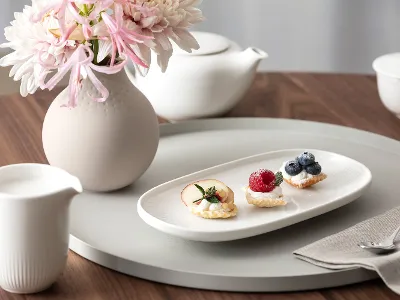 Piccole delizie dolci con Afina di Villeroy & Boch Un piatto bianco Afina con tre piccoli pasticcini e frutta si trova su un vassoio rotondo accanto a un vaso di fiori, una teiera e un brocchetto di latte su un tavolo di legno.
