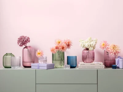 Szklane wazony Fleur w różnych pastelowych kolorach i rozmiarach Różne szklane wazony w pastelowych kolorach z kolekcji Fleur, ozdobione kwiatami, są ułożone na jasnej powierzchni z książkami i małymi przedmiotami dekoracyjnymi przed różową ścianą.