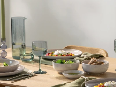 A modern dining table with a carafe, gray tableware, glasses, food, and green napkins in natural light.
