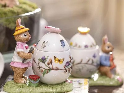 Niedliche Porzellanfigur Osterhäsin Anna gibt dem großen Osterei die letzten Pinselstriche, während der süße Osterhase Max an seiner Karotte knabbert und sich an das große Osterei-Porzellangefäß lehnt