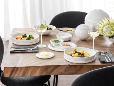 A table set with La Boule and black and gray chairs around it A table set with La Boule with two plates full of food, two glasses of white wine, various bowls with side dishes, and a flower arrangement with black and gray chairs around it.