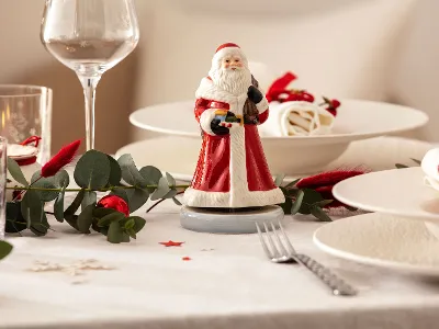 Une table de fête dressée avec une boîte à musique en forme de Père Noël Nostalgic Melody Une table festive avec une boîte à musique en forme de Père Noël Nostalgic Melody au centre, entourée d’assiettes, de verres, de couverts de table et de plantes vertes.