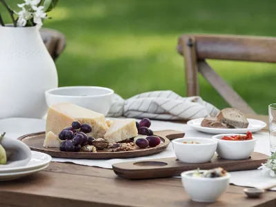 Un élégant couvert Artesano de Villeroy & Boch se compose d'une table en bois décorée de fromage, de raisins, de noix et de pain. Un élégant couvert Artesano de Villeroy & Boch se compose d'une table en bois décorée de fromage, de raisins, de noix et de pain. Des coupes en céramique stylées et un vase blanc avec des fleurs rehaussent le décor rustique.