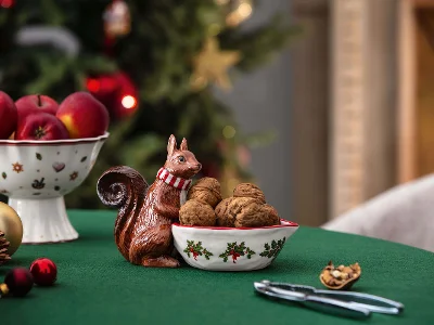 Toy's Fantasy bowl with walnuts on a Christmas table A Toy's Fantasy bowl decorated with a squirrel figurine with walnuts next to a nutcracker on a green tablecloth. Next to it is a bowl of red apples, with a decorated Christmas tree in the background.