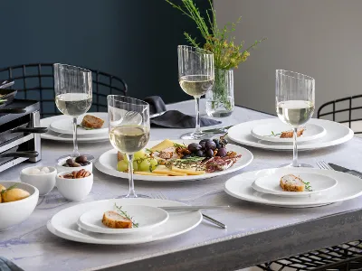 Ein gedeckter Esstisch, geschmückt mit der eleganten NewMoon-Kollektion von Villeroy & Boch, mit vier Gedecken, zu denen jeweils ein Glas Weißwein gereicht wird. In der Mitte steht eine Platte mit Käse, Trauben und Feigen, umgeben von Schalen mit Kartoffeln, Nüssen und Oliven.