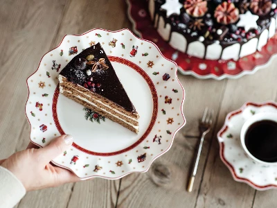 On the table there is a decorated complete cake, a cup of coffee, a fork and a saucer and plate set from Toy's Delight. A hand holding a plate with a slice of chocolate cake. On the table there is also a decorated complete cake, a cup of coffee, a fork and a saucer and plate set from Toy's Delight.