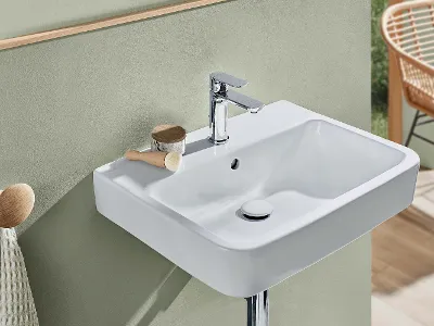 Weißes O.novo-Waschbecken mit verchromter Armatur Ein modernes rechteckiges weißes O.novo-Waschbecken mit verchromtem Wasserhahn, Bürste und kleinem Behälter an der Rückwand vor einer mattgrünen Wand. Im Hintergrund steht ein beiger Stuhl.