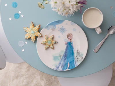 Une assiette avec une figurine Frozen, deux biscuits en forme de flocon de neige, une tasse de lait et une cuillère sur une table bleue.