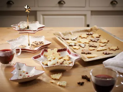 Ein Holztisch mit sternförmigen Schalen von Winter Bakery Delight Ein Holztisch mit sternförmigen Schalen von Villeroy & Boch Winter Bakery Delight, auf denen dekorierte Kekse liegen, und einem Backblech mit festlichen Keksen auf Backpapier. Auf dem Tisch stehen zwei Gläser mit rotem Getränk.