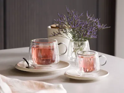 Artesano-Glasbecher gefüllt mit rosa Tee, auf Untertassen neben einem Löffel auf einem Tisch platziert. Artesano-Glasbecher von Villeroy & Boch, gefüllt mit rosa Tee, auf Untertassen neben einem Löffel auf einem Tisch platziert, der mit einer kleinen Vase mit lila Blumen geschmückt ist.
