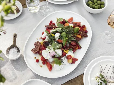 Piatto Artesano con insalata mista Su una tovaglia bianca è presente un piatto Artesano di Villeroy & Boch con un'insalata mista di pomodori, erbe e salumi. Accanto ci sono ciotole con olive, un bicchiere di rosé e una ciotola con del sale.