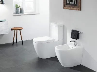 Un WC Subway 2.0 à côté d’un bidet sur un sol carrelé gris Une salle de bains moderne avec des toilettes blanches et un bidet sur un sol carrelé. Sur le côté gauche, il y a un tabouret en bois et une petite armoire, et sur le mur, une fenêtre et une œuvre d’art encadrée.
