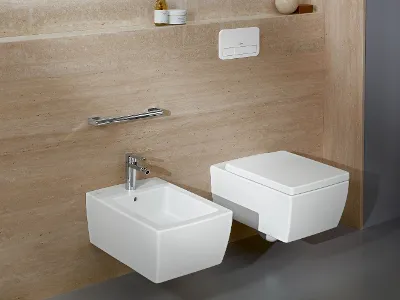 Weiße Toilette und Bidet aus der Kollektion Memento 2.0 Modernes Bad mit weißem Wand-WC und Bidet vor beige gefliesten Wänden, ausgestattet mit verchromter Armatur, Toilettenspülung und elegantem Handtuchhalter.