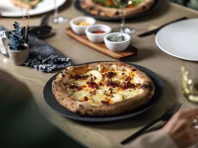 Une assiette noire Manufacture avec de la pizza fraîchement sortie du four Une pizza fraîchement cuite sur une assiette noire Manufacture, accompagnée de différentes épices et d’un verre de vin blanc.