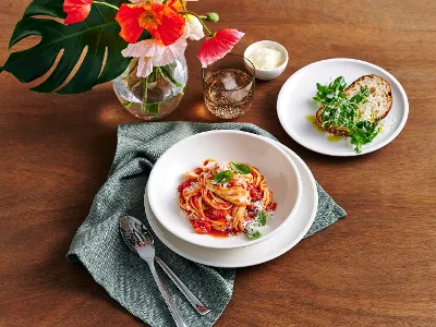 Artesano dinner set for four, arranged on a rustic wooden table, features a delicious spread of pasta and fresh bread. Accented with a blue linen napkin and silver cutlery, the setting is further elevated with elegant Like glassware and a vase filled with florals.