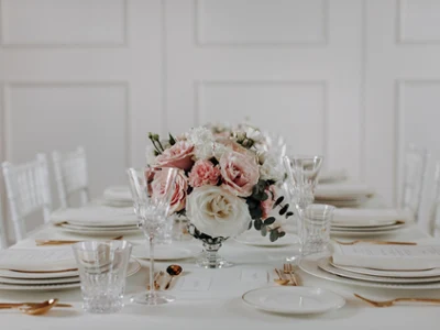 Elegant dining table set with white plates, crystal glasses, and gold cutlery. A centerpiece of pink and white flowers adds a romantic touch.