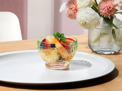 One Boston bowl by Villeroy & Boch with a pink base are placed on a tray. One Boston bowl with fruits in it by Villeroy & Boch with a pink base are placed on a tray next to a Fleur vase with white and pink flowers.