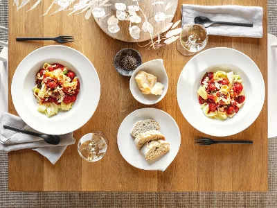 Flow Pastateller mit Nudeln auf einem Tisch für 2 Personen Zwei Flow-Teller mit Pasta, Tomaten und Käse von Villeroy & Boch auf einem Holztisch, begleitet von einer Schüssel Brot, zwei Weingläsern, Besteck und einem dekorativen Tafelaufsatz.