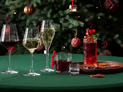 Festliche Toys Delight Gläser vor einem geschmückten Weihnachtsbaum Ein grüner Tisch mit fünf verschiedenen "Toy´s Delight" Gläsern von Villeroy & Boch, einem Holzbrett mit getrockneten Orangenscheiben und einem geschmückten Weihnachtsbaum im Hintergrund.