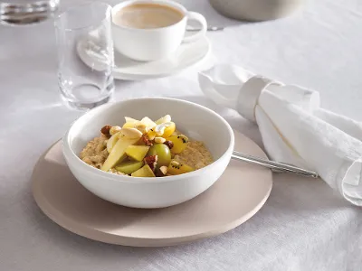 Ciotola per cereali NewMoon con porridge Una ciotola NewMoon con del porridge, guarnita con frutta a fette e noci, su un tavolo, con un bicchiere d'acqua e una tazza di caffè. Sono presenti anche una candela accesa e un tovagliolo piegato.
