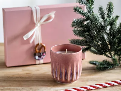 Une bougie parfumée Perlemor rose est posée sur une surface en bois à côté d’une petite branche de sapin, d’une boîte cadeau rose avec un nœud blanc et un ornement ainsi que d’un grand sucre d’orge rayé.