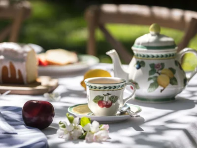 French Garden coffee mug on a white tablecloth Outdoor table setting with tea and snacks, consisting of a patterned teapot, teacup, cake, and fruit in natural daylight.