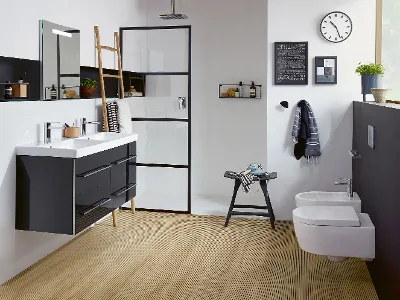 Badezimmer der Kollektion Avento von Villeroy & Boch Modernes Badezimmer mit Doppelwaschbecken, schwarz gerahmter Dusche, Holzboden, wandmontiertem WC und Bidet, einer Uhr, einem Leiterregal und verschiedenen Dekorationsgegenständen.