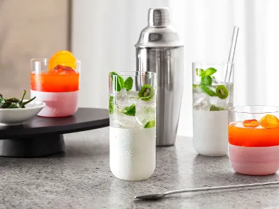 White Manufacture glasses with cocktails Four colorful cocktails in white Manufacture glasses on a marble surface, a cocktail shaker in the background, and a small bowl of bell peppers on a tray.