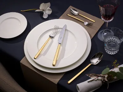 La Classika Geschirr mit goldenen Akzenten auf weißen Tellern Kreieren Sie eine elegante Tischdekoration mit der Kollektion La Classica von Villeroy & Boch, komplett mit weißem Teller, goldenem Besteck, Weinglas, Wasserglas, Serviette und einer dekorativen Orchidee auf einer dunklen Tischdecke.