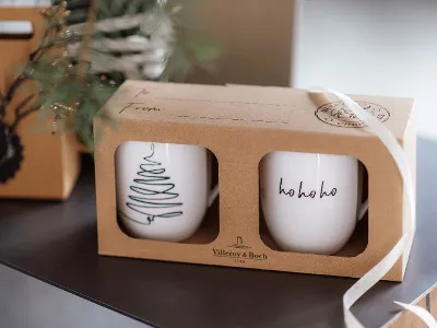 Two Statement cups with a Christmas pattern in a cardboard box as a gift Two Statement cups in a cardboard box, one with a Christmas tree pattern and the other with the text "ho ho ho". The box is partly wrapped in a white bow.